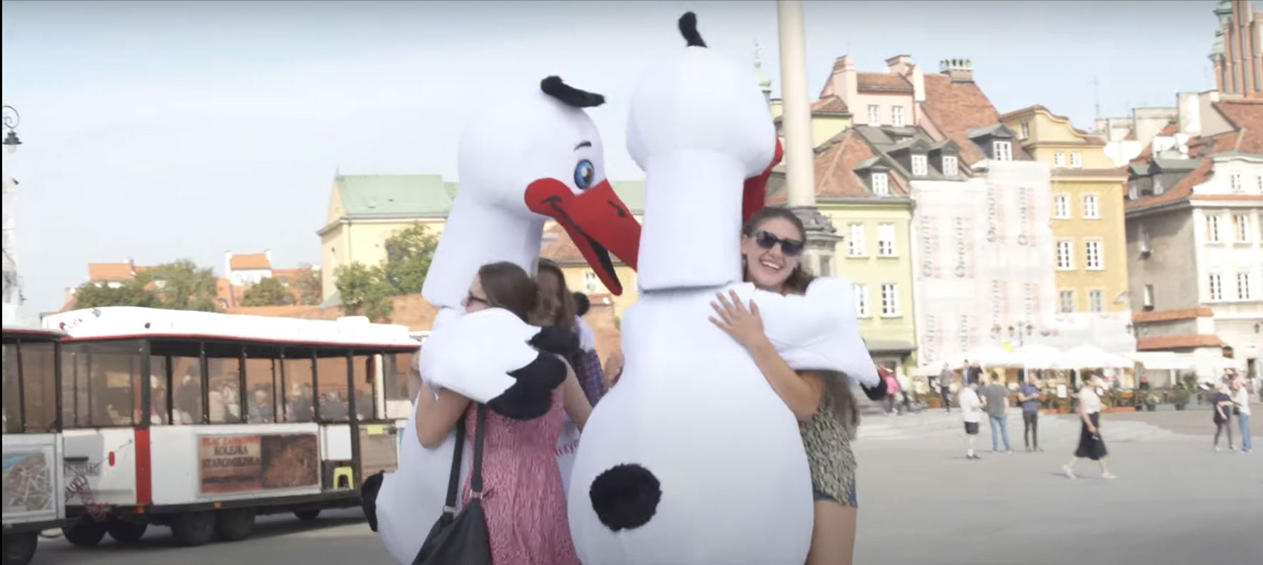 Na fotografii widnieją dwa stroje w postaci bociana w objęciach dwóch młodych kobiet na Placu Zamkowym w Warszawie